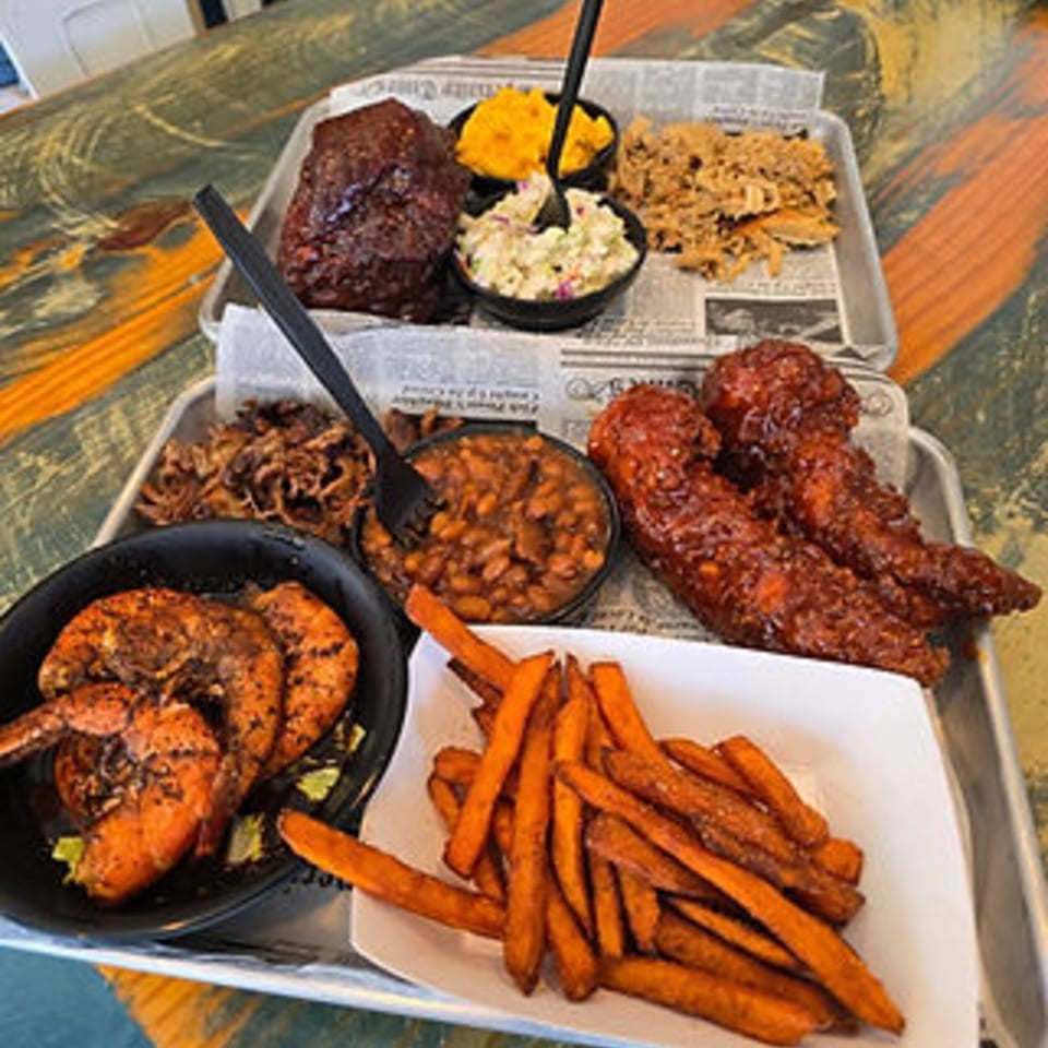 A metal tray filled with various BBQ dishes, including ribs, pulled pork, shrimp, baked beans, and sweet potato fries.