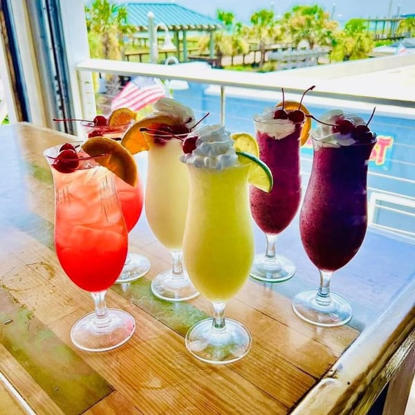 Six colorful tropical drinks with whipped cream and fruit garnishes on a wooden bar overlooking the ocean.