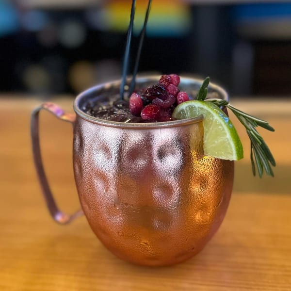 A copper mug cocktail with berries, a lime wedge, and rosemary sprig on a wooden table.