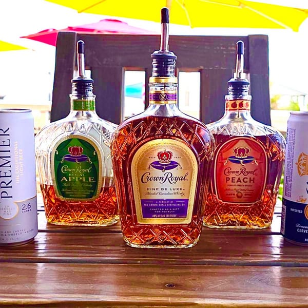 Three bottles of Crown Royal whiskey, Apple, Fine De Luxe, and Peach, on a wooden table.