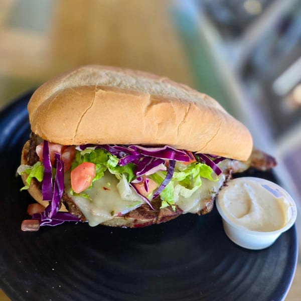 A BBQ sandwich with melted cheese, lettuce, red cabbage, and tomatoes, with a side of sauce.