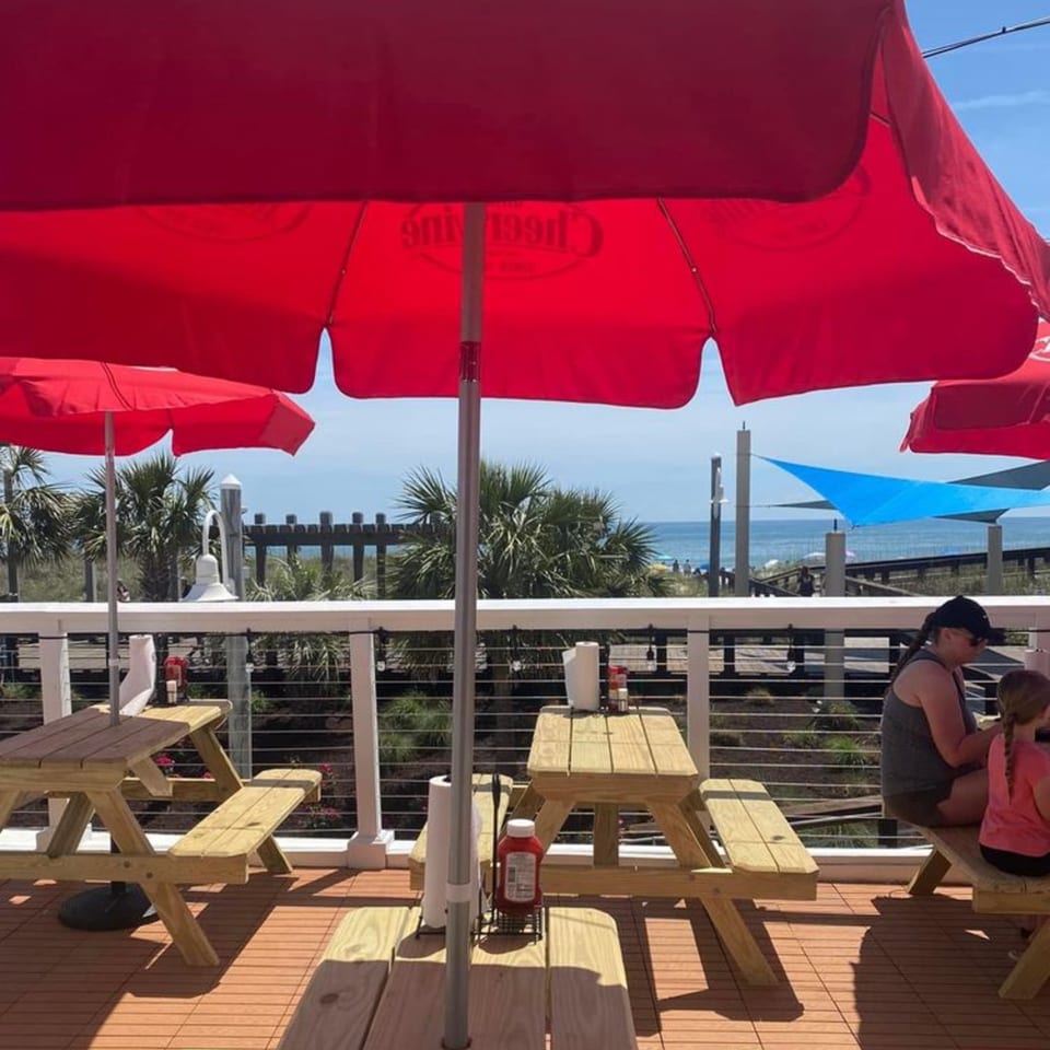 Sunny Daze Smokehouse's outdoor patio with red umbrellas, picnic tables, and ocean views.