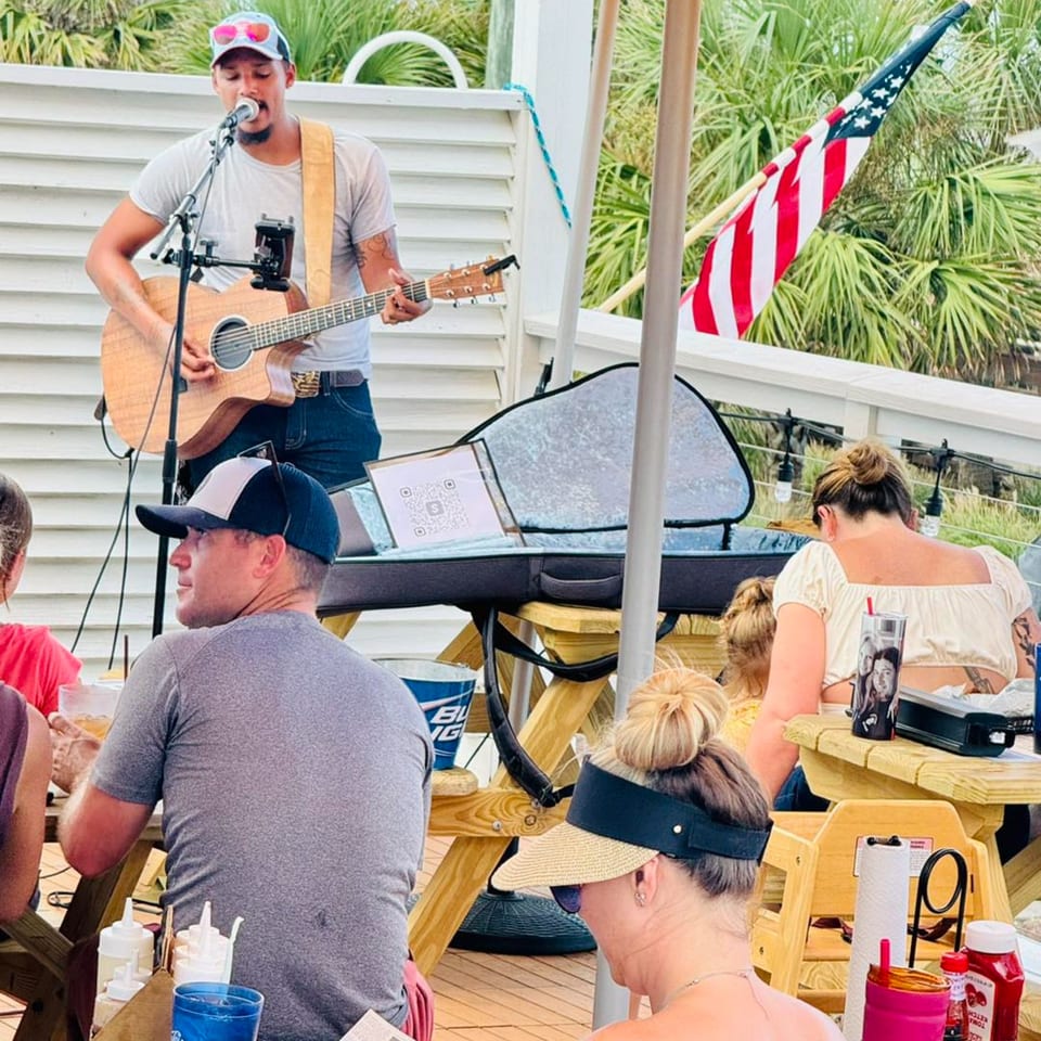 A musician plays guitar and sings for patrons seated at picnic tables on an outdoor patio.