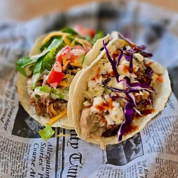 Two Sunny Daze Smokehouse BBQ tacos with various toppings on newspaper-patterned paper.