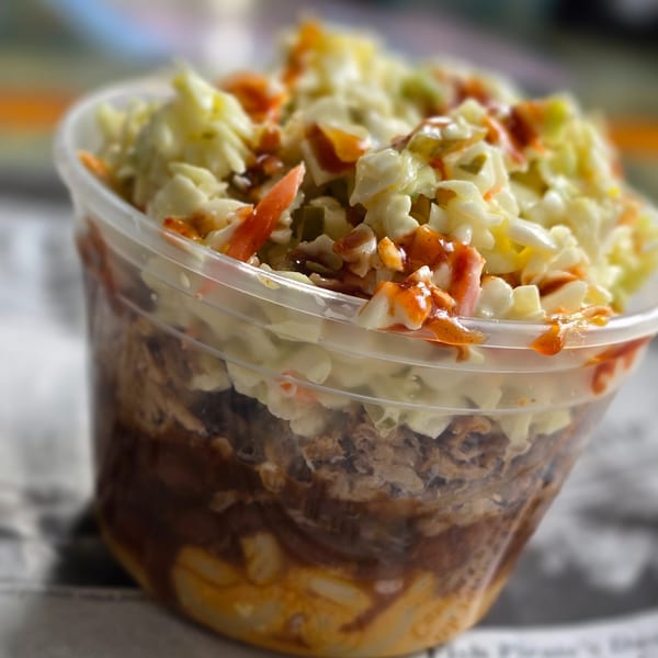 A clear plastic cup filled with layers of BBQ pulled pork, macaroni and cheese, and coleslaw.