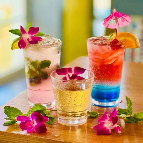 Three colorful cocktails, garnished with orchids, mint, lime, and an umbrella, on a wooden surface.