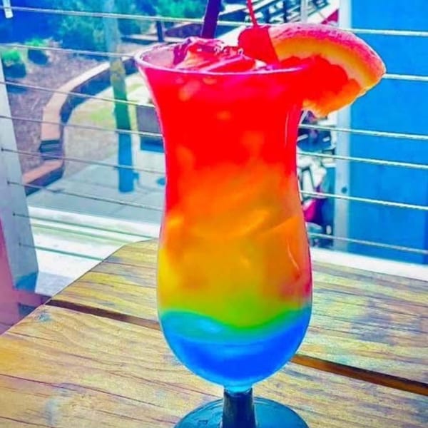 A vibrant, rainbow-layered cocktail with a cherry and orange slice on a wooden table.