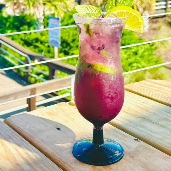 A vibrant purple cocktail with mint and lime sits on a wooden table outdoors at Sunny Daze Smokehouse.