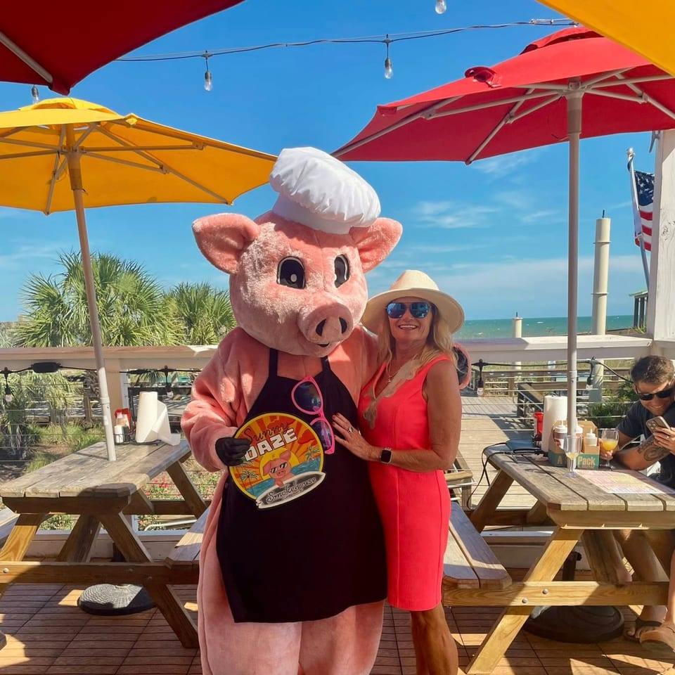 A woman in a red dress poses with the Sunny Daze Smokehouse pig mascot on an outdoor patio.