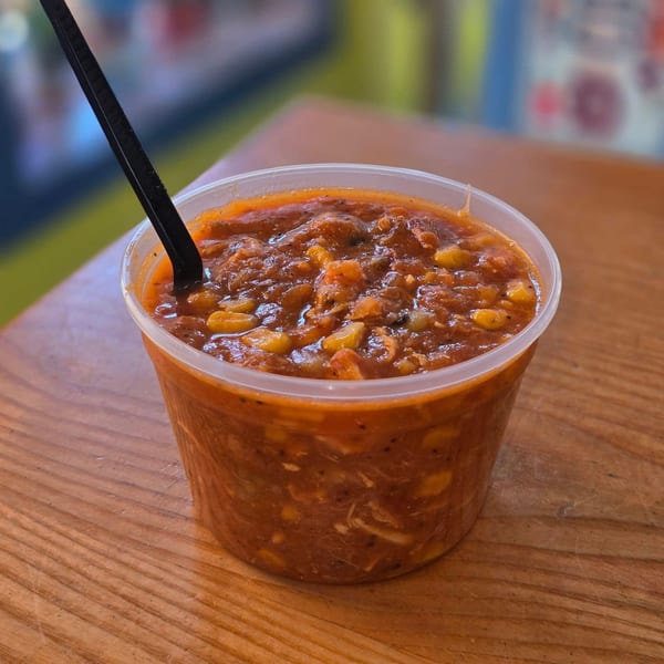 A clear plastic container of Sunny Daze Smokehouse baked beans with a black spoon on a wooden table.