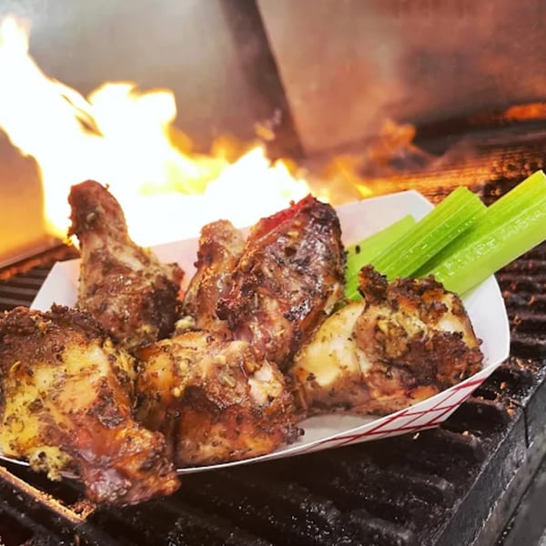 Smoked chicken wings and drumsticks with celery in a paper tray, with flames in the background.