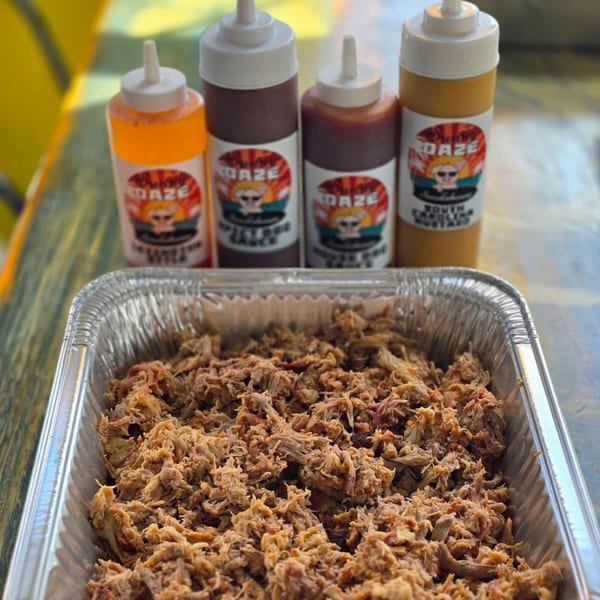A foil pan of pulled pork with four Sunny Daze Smokehouse BBQ sauce bottles in the background.