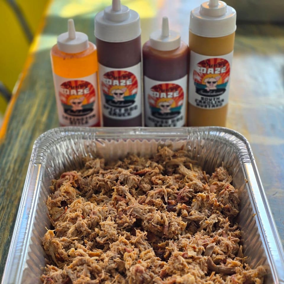 A foil pan of pulled pork with four Sunny Daze Smokehouse BBQ sauce bottles in the background.