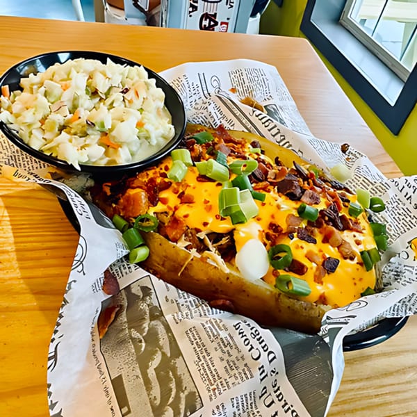 A loaded baked potato with cheese, bacon, and green onions, served with a side of coleslaw.
